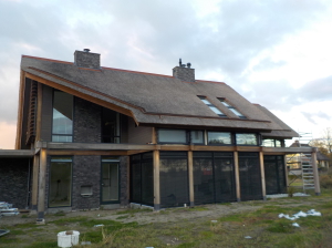 Luxe woning architectuur met natuurlijk materiaalgebruik, chique rieten dak afgedekt met strakke nokvorst van Cortenstaal, Fraké houten Post & Beam structuur voorzien van Luxaflex buitenzonwering.